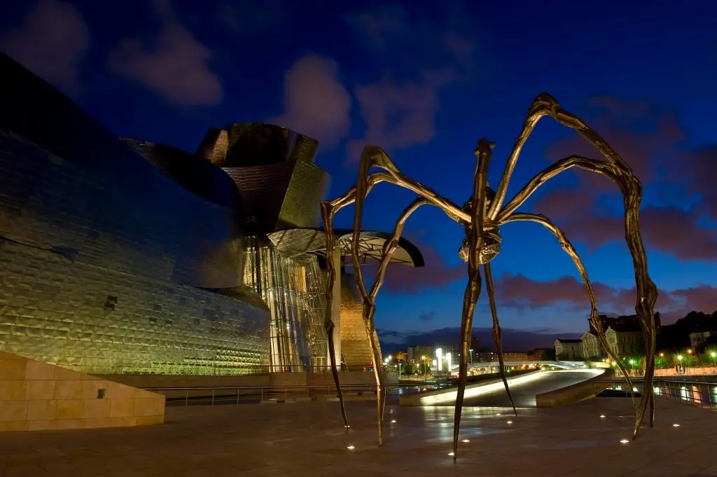 Il ragno Maman illuminato al crepuscolo davanti al museo Guggenheim di Bilbao, con il cielo blu intenso e le luci della città sullo sfondo